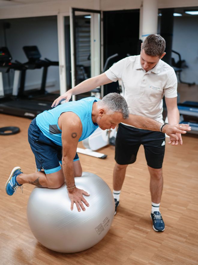 Physiotherapeut unterstützt einen Mann beim Gleichgewichtstraining auf einem Gymnastikball im Fitnessraum.