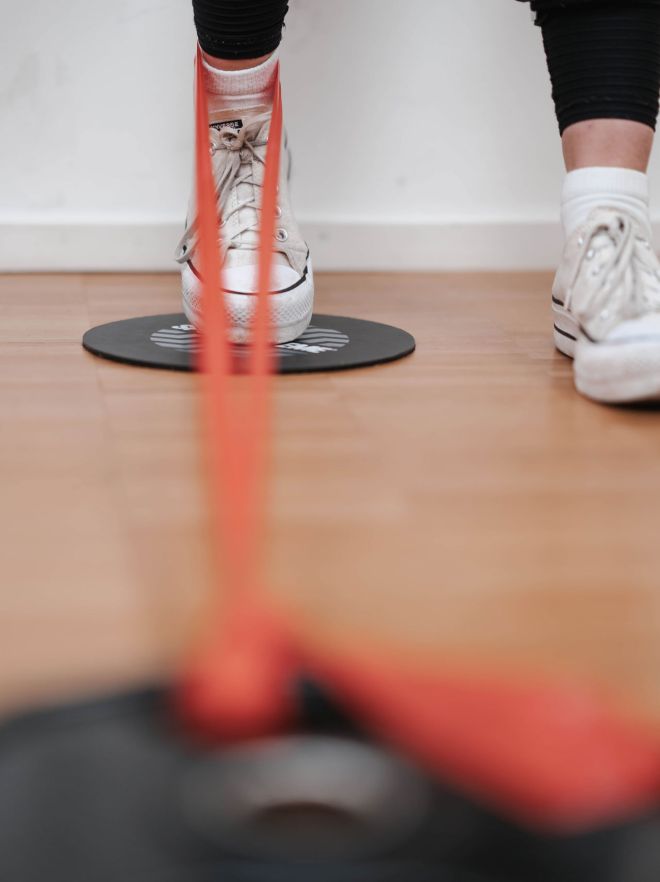 Nahaufnahme eines Fußes mit Turnschuh, der mit einem roten Widerstandsband auf einer Balance-Scheibe trainiert.